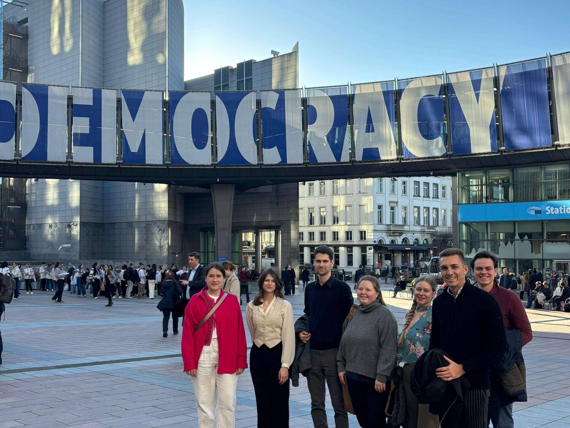 Eine Gruppe junger Menschen steht auf dem Platz vor dem EU-Parlament in Brüssel. Im Hintergrund ein Banner mit der Aufschrift Democracy.