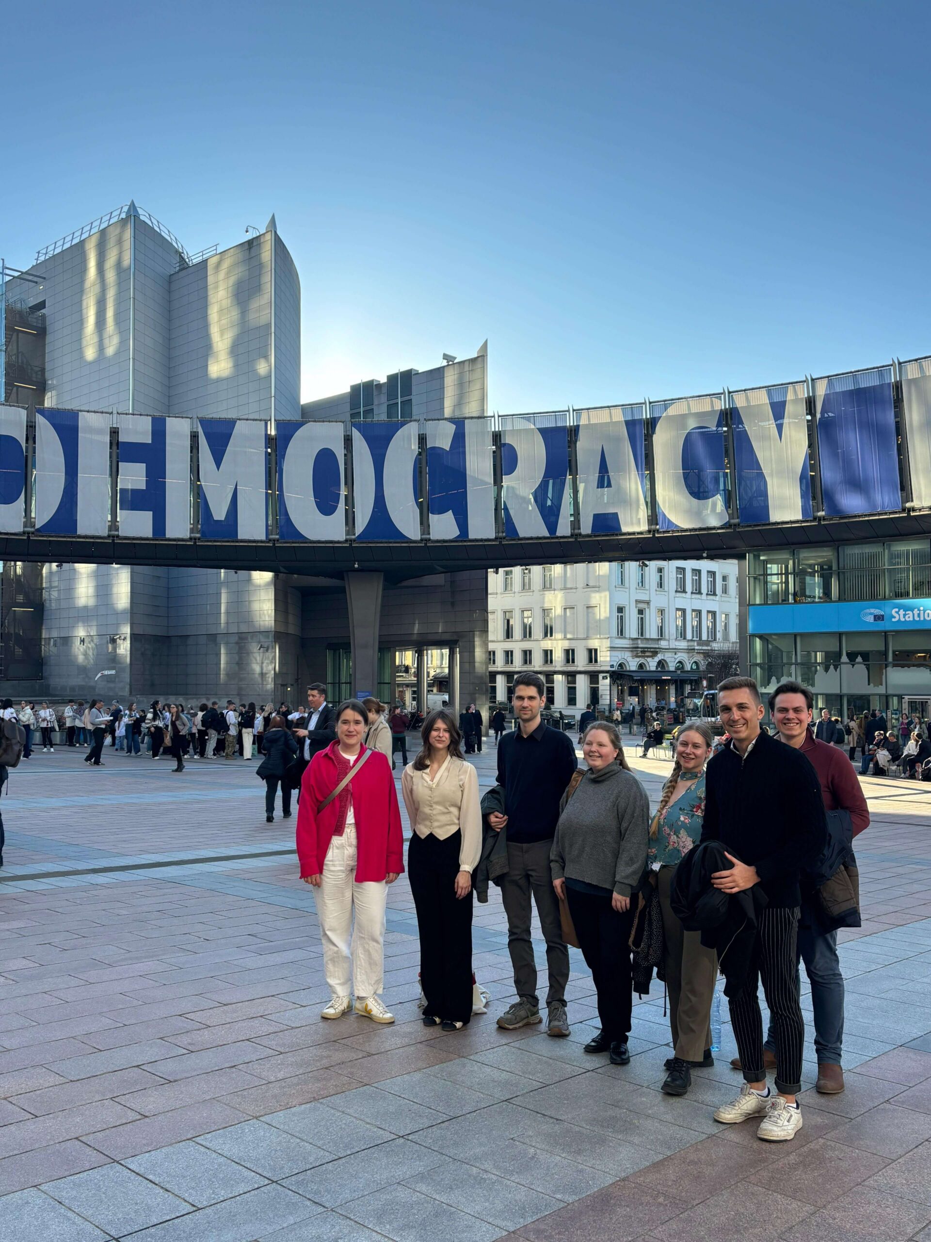 Eine Gruppe junger Menschen steht auf dem Platz vor dem EU-Parlament in Brüssel. Im Hintergrund ein Banner mit der Aufschrift Democracy.