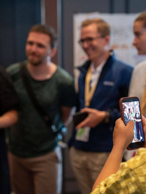 Im Hintergrund des Bildes sprechen junge Menschen miteinander. Die Szene wird von einer Person im Vordergrund mit einem Smartphone aufgenommen.