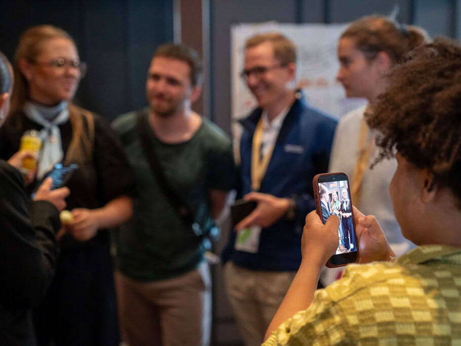 Im Hintergrund des Bildes sprechen junge Menschen miteinander. Die Szene wird von einer Person im Vordergrund mit einem Smartphone aufgenommen.
