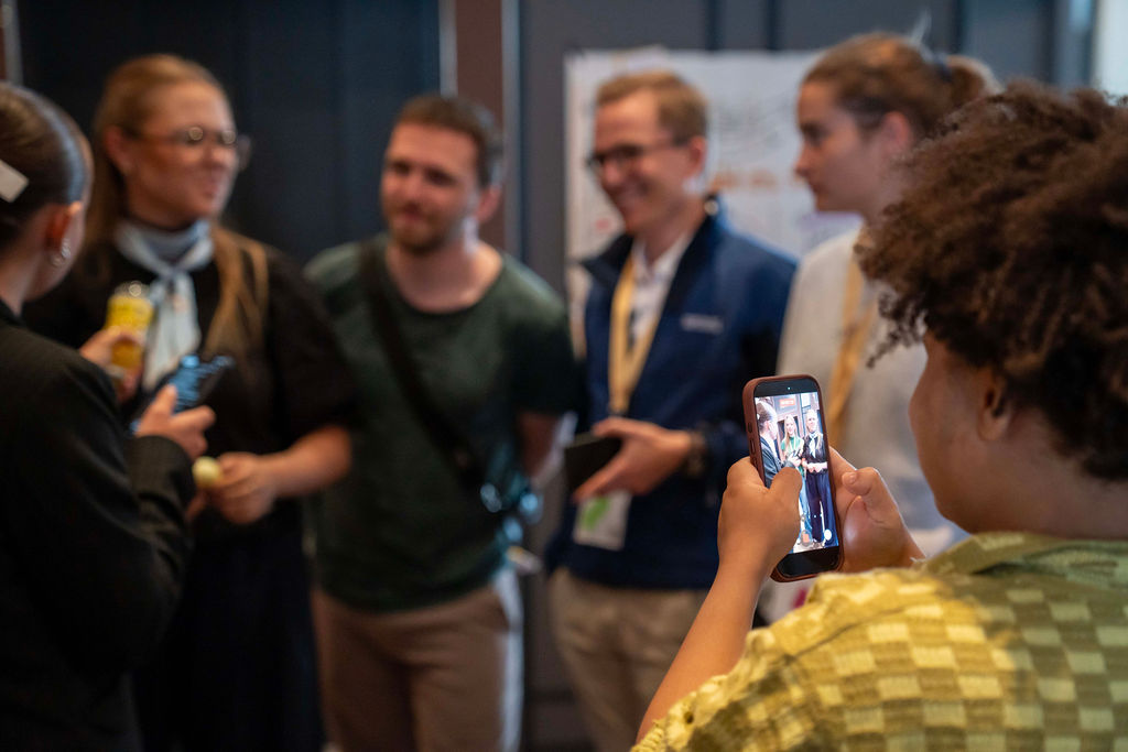 Im Hintergrund des Bildes sprechen junge Menschen miteinander. Die Szene wird von einer Person im Vordergrund mit einem Smartphone aufgenommen.